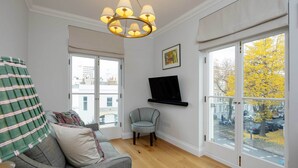 Ein Wohnzimmer mit grauem Sofa, einem kleinen Sessel, einem Fernseher an der Wand und großen Fenstern mit Außenblick.