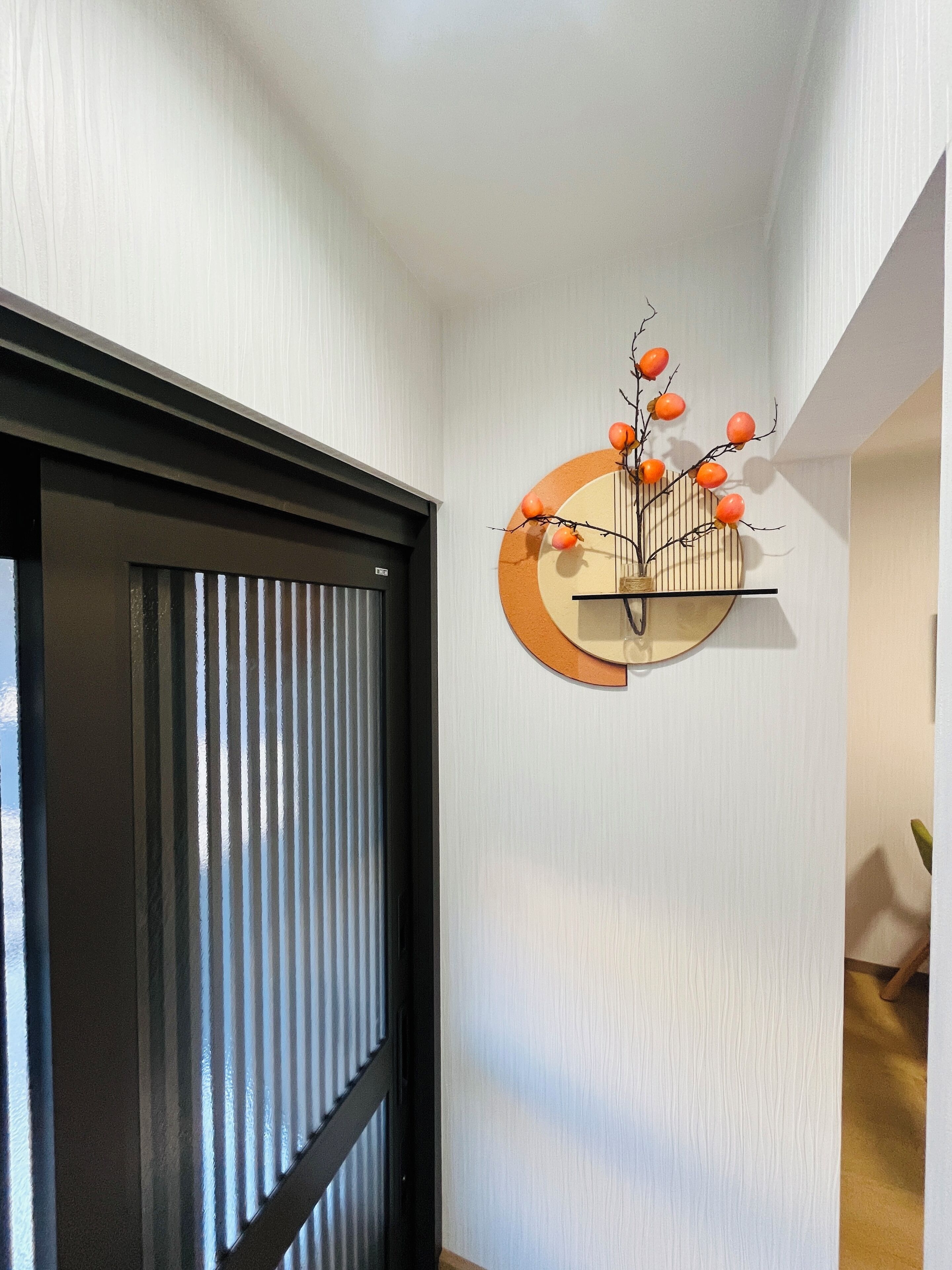 A hallway with a black-framed door, a wall-mounted decorative piece with branches and orange spheres, and a wooden floor.