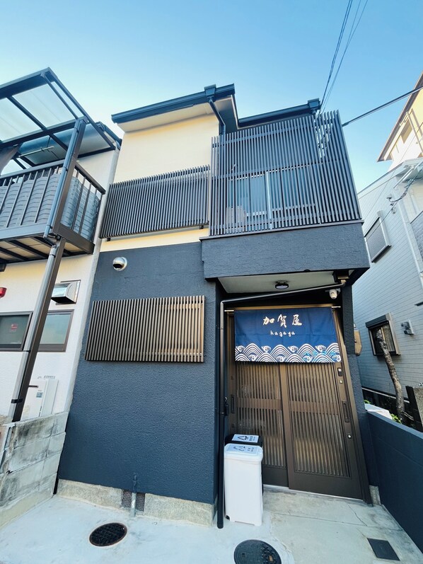 Un edificio de fachada oscura, con un balcón con barandilla metálica y una entrada azul con caracteres japoneses.