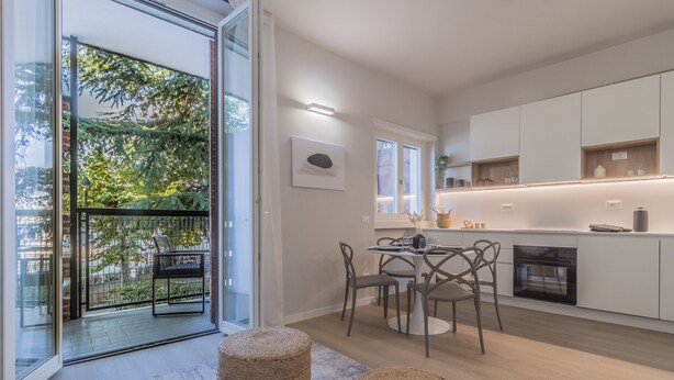 Una cocina moderna con armarios blancos, un comedor con mesa y sillas, y un balcón con vistas a árboles.