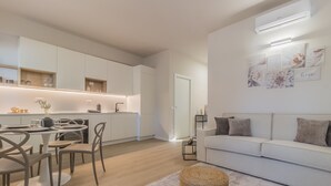 Una sala de estar moderna con un sofá blanco, una mesa de comedor con sillas y una cocina con armarios blancos y horno empotrado.