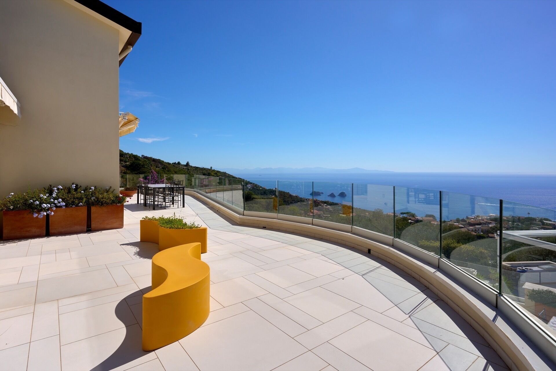 Un balcón con un banco amarillo curvo, una mesa con sillas y vistas al mar y montañas lejanas.