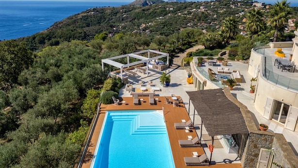 Zona de piscina en la azotea con sillones de descanso y una pérgola, con vistas a un paisaje costero con casas y vegetación.