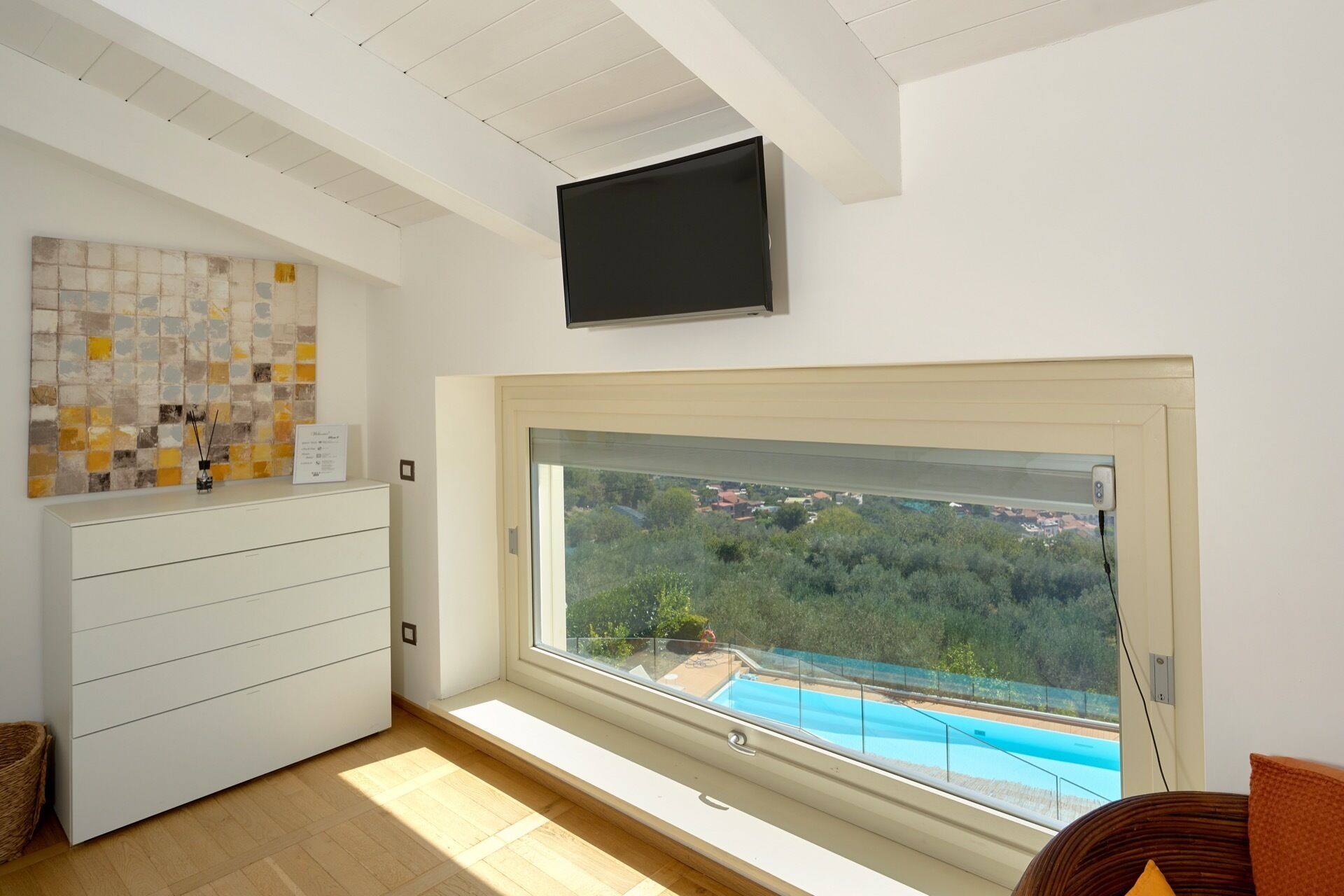 Une chambre moderne dotée d’une grande fenêtre donnant sur une piscine et un espace verdoyant, d’un téléviseur à écran plat fixé au-dessus de la fenêtre, et d’une commode blanche au design minimaliste agrémentée d’un pare-feu en carreaux décoratifs.