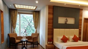 A hotel room with a wooden floor, a bed with white and orange bedding, a small table with chairs, and a window with curtains. - Hotel yelchiko (Varanasi)