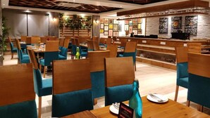 A restaurant with wooden tables and blue chairs, a bar area with chalkboard menus, and framed pictures on the walls. - Hotel yelchiko (Varanasi)