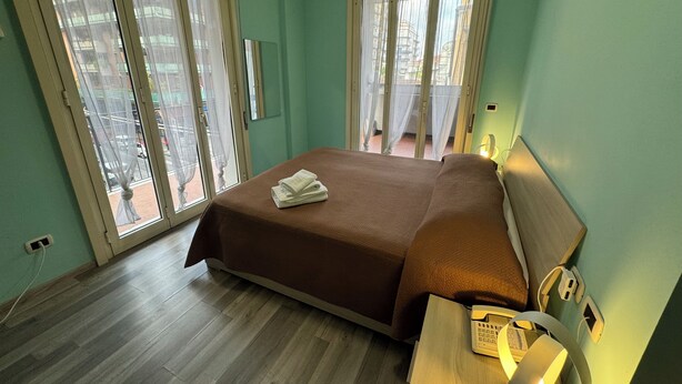 Ein Schlafzimmer mit einem Bett, einem Nachttisch mit Lampe, einem Telefon und einem Fenster mit Vorhängen.