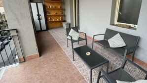 Ein Balkon mit einem gemusterten Boden, zwei schwarze Metallstühle mit Kissen, ein kleiner schwarzer Tisch und ein Holzregal mit diversen Gegenständen.