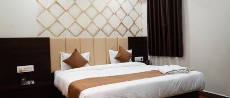 A hotel room with a large bed, a headboard with a diamond pattern, a nightstand with a lamp, and a window with curtains.