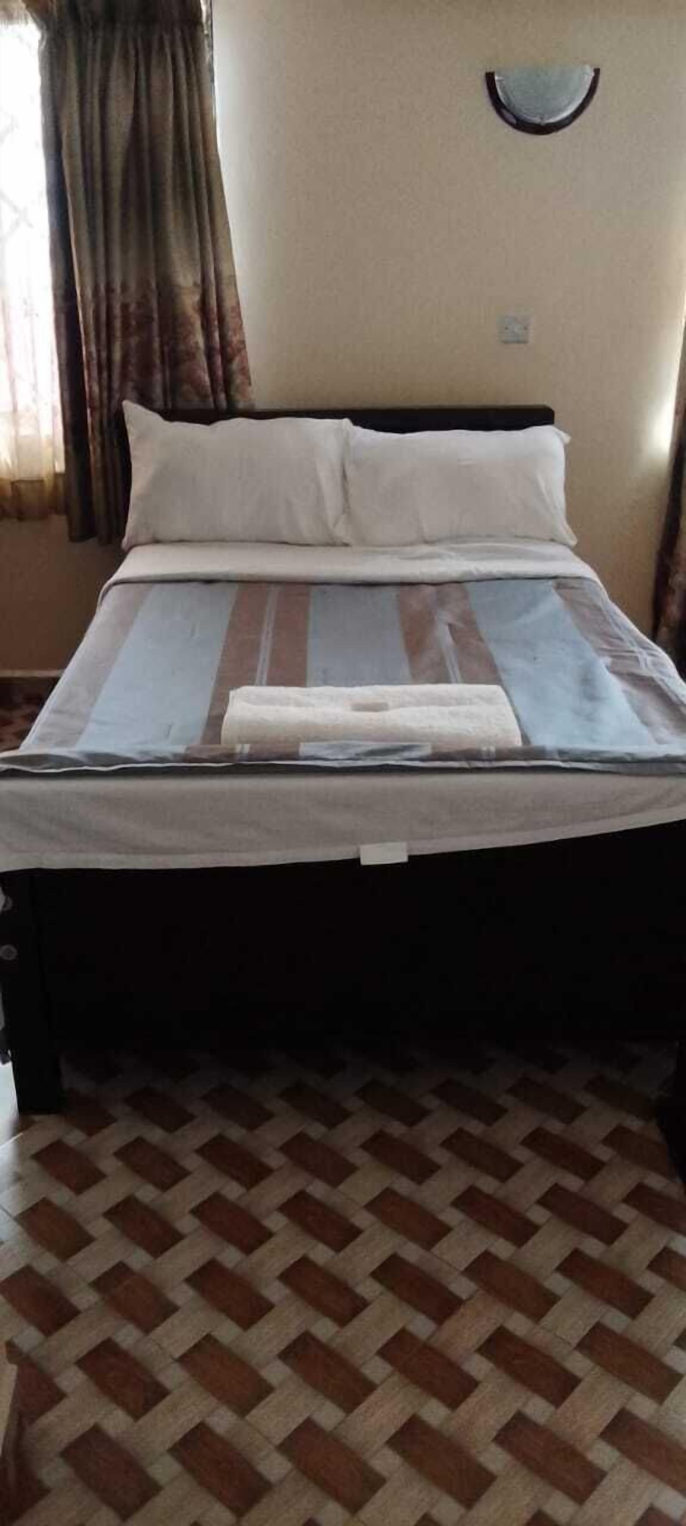 Una cama con una colcha a rayas, dos almohadas y una manta doblada. La habitación tiene un suelo de cuadros y una ventana con cortinas.