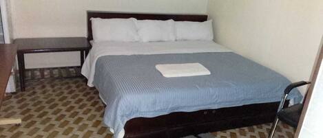 A single bed with a wooden frame, a blue bedspread, and white pillows. There is a small bedside table and a chair to the left of the bed.