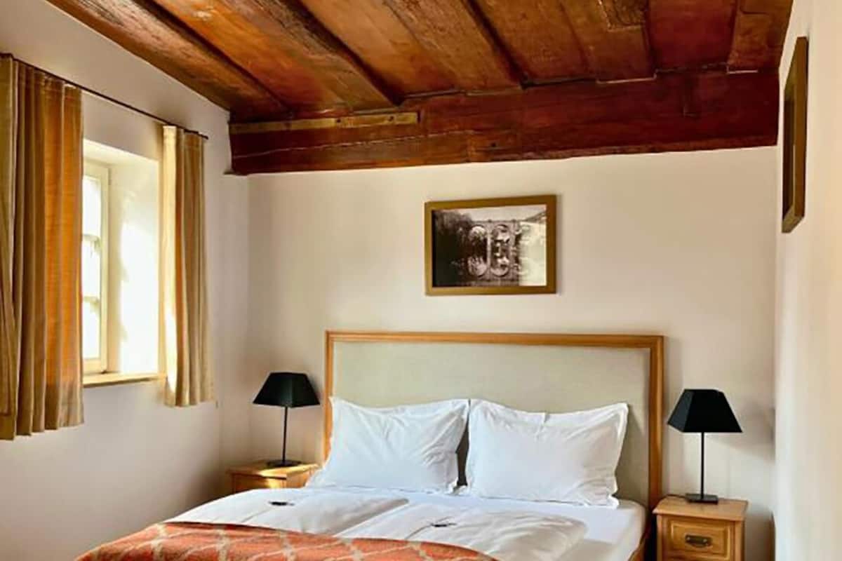 A bedroom with a wooden ceiling, a bed with a patterned bedspread, two bedside tables with lamps, and a framed picture on the wall.