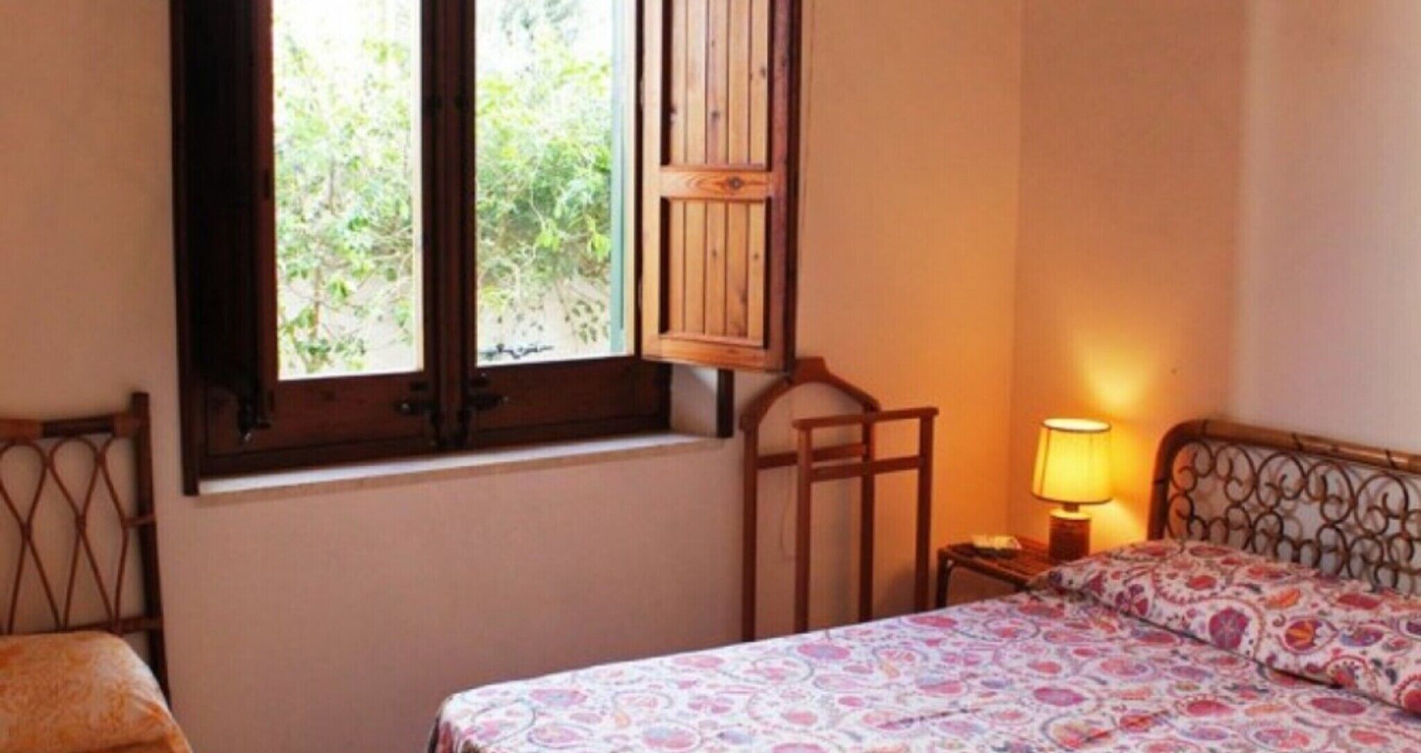 Un dormitorio con cama, ventana con contraventanas de madera, mesita de noche con lámpara y una silla.