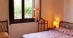 A bedroom with a bed, a window with wooden shutters, a nightstand with a lamp, and a chair. - Villetta Sole: Holiday Home with Mountain View, Shared Terrace & Air Conditioning (San Vito Lo Capo)