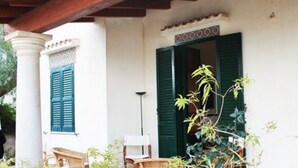 A building with a white exterior, green shutters, and a covered porch with wicker furniture. - Villetta Sole: Holiday Home with Mountain View, Shared Terrace & Air Conditioning (San Vito Lo Capo)