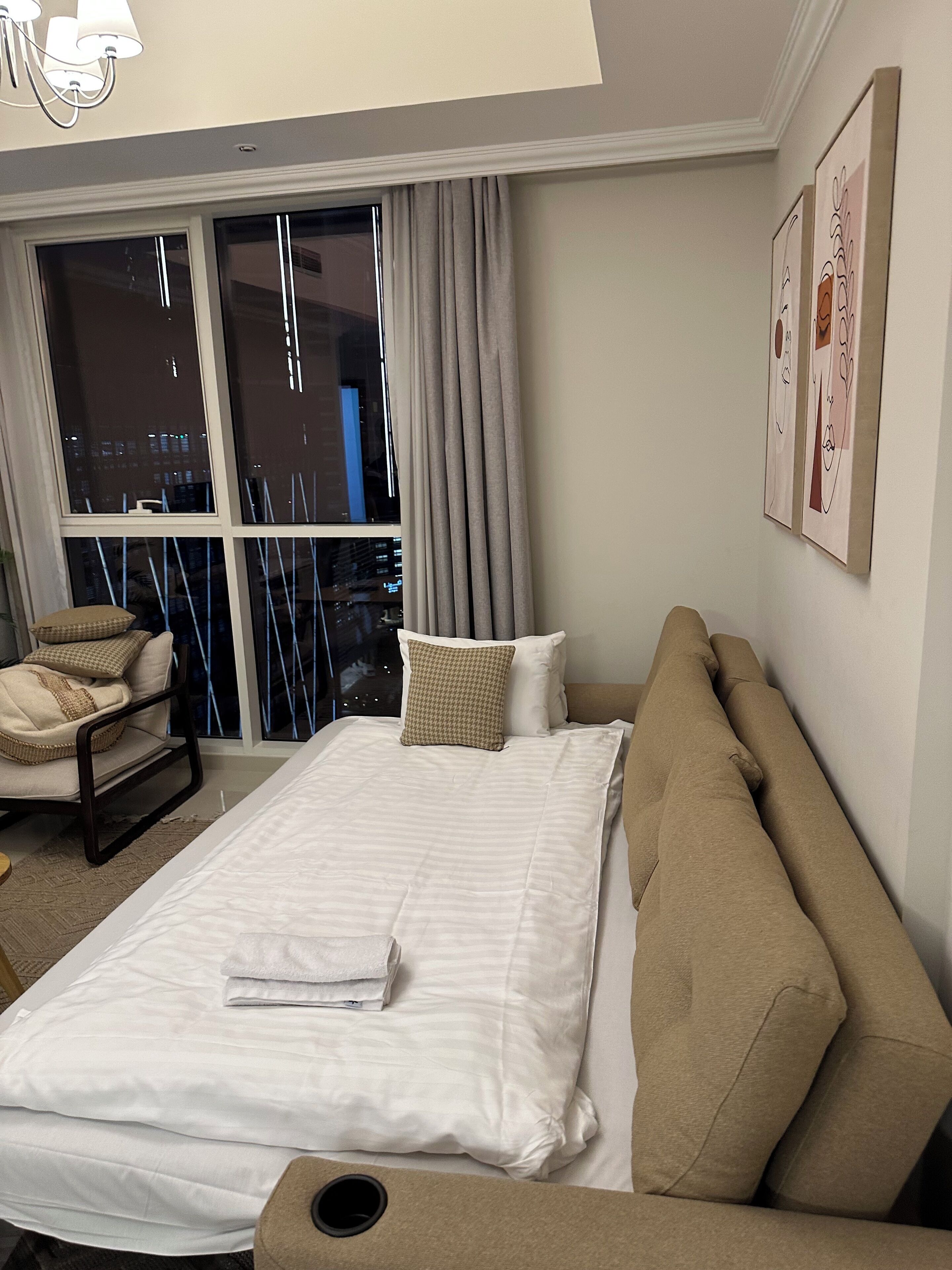 Une chambre d’hôtel avec un grand lit, un canapé, un fauteuil et une vue sur la ville la nuit, visible par la fenêtre.