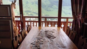 Une table à manger en bois, ornée de motifs sculptés, placée dans une pièce dotée de grandes fenêtres donnant sur un paysage montagneux.