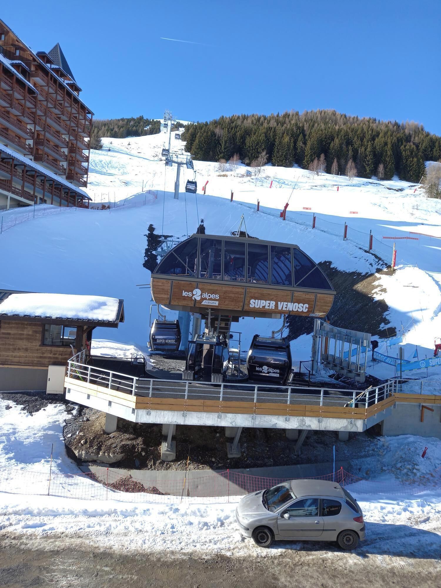 Eine Talstation einer Seilbahn mit einem Holzbauten und einer Glasunterstand, auf dem "les Alpes AERON SUPER VENDO SC" steht.