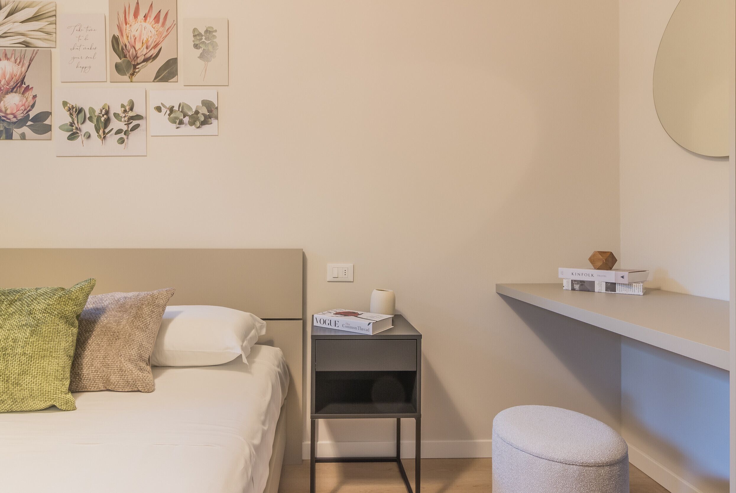 A bedroom with a bed, pillows, a nightstand with a book and a lamp, a small stool, and a wall with framed botanical prints.