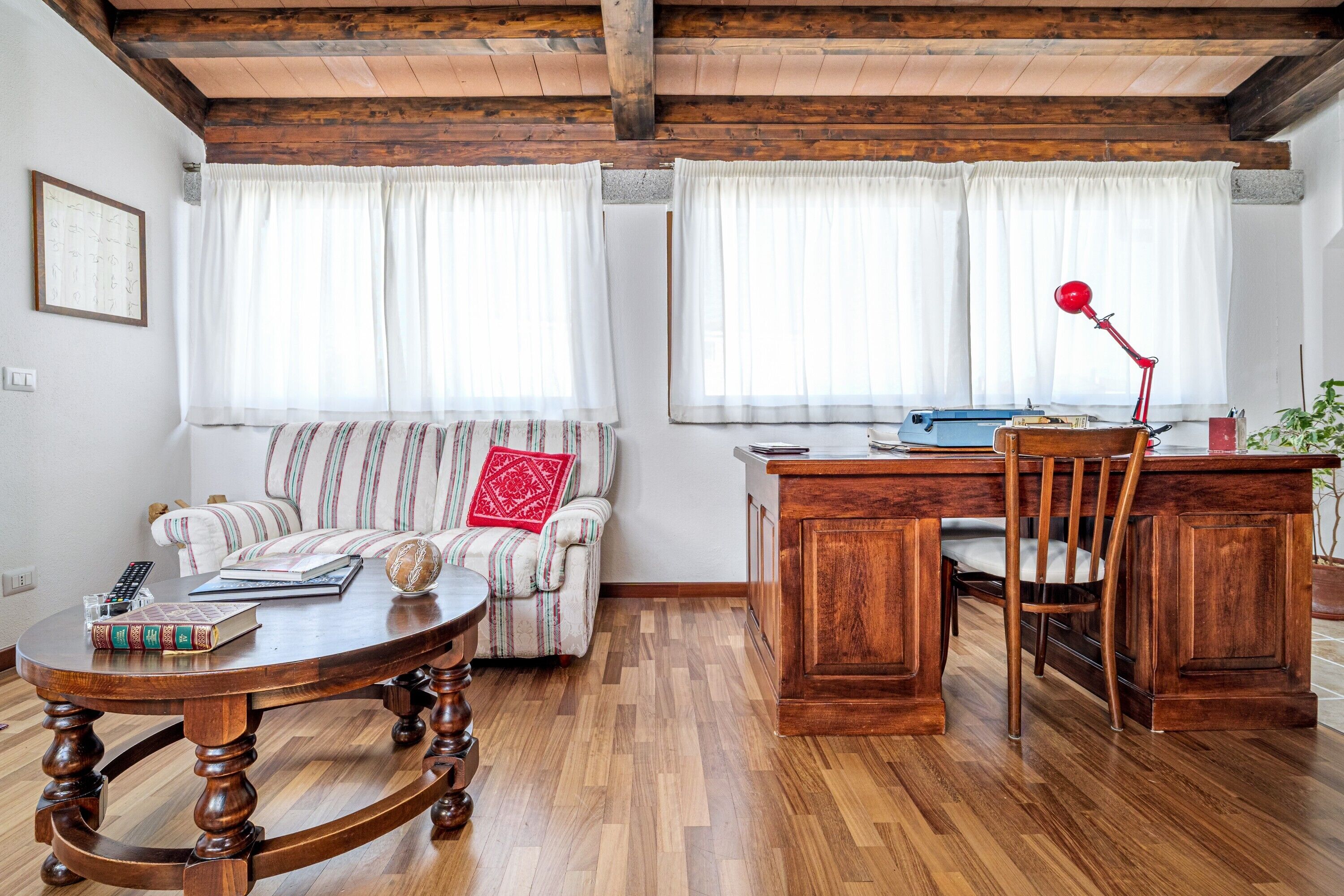 A living room with a wooden table, a sofa, a desk with a chair, and a red lamp.