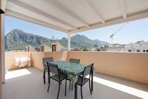 Een overdekt buitengebied met een tafel en stoelen, een uitzicht op bergen en een bouw kraan in de verte.