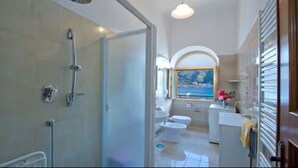 Une salle de bain avec douche, toilettes et lavabo, donnant sur la mer grâce à une fenêtre en arc de cercle.