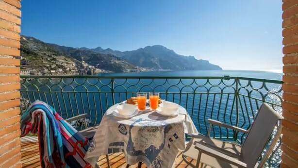 Un balcon avec une table préparée pour le petit-déjeuner, offrant une vue sur un paysage côtier avec des montagnes et la mer.