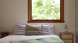 Ein Schlafzimmer mit Bett, Kissen, einer Nachttischlampe und einem Fenster mit Blick auf Bäume.