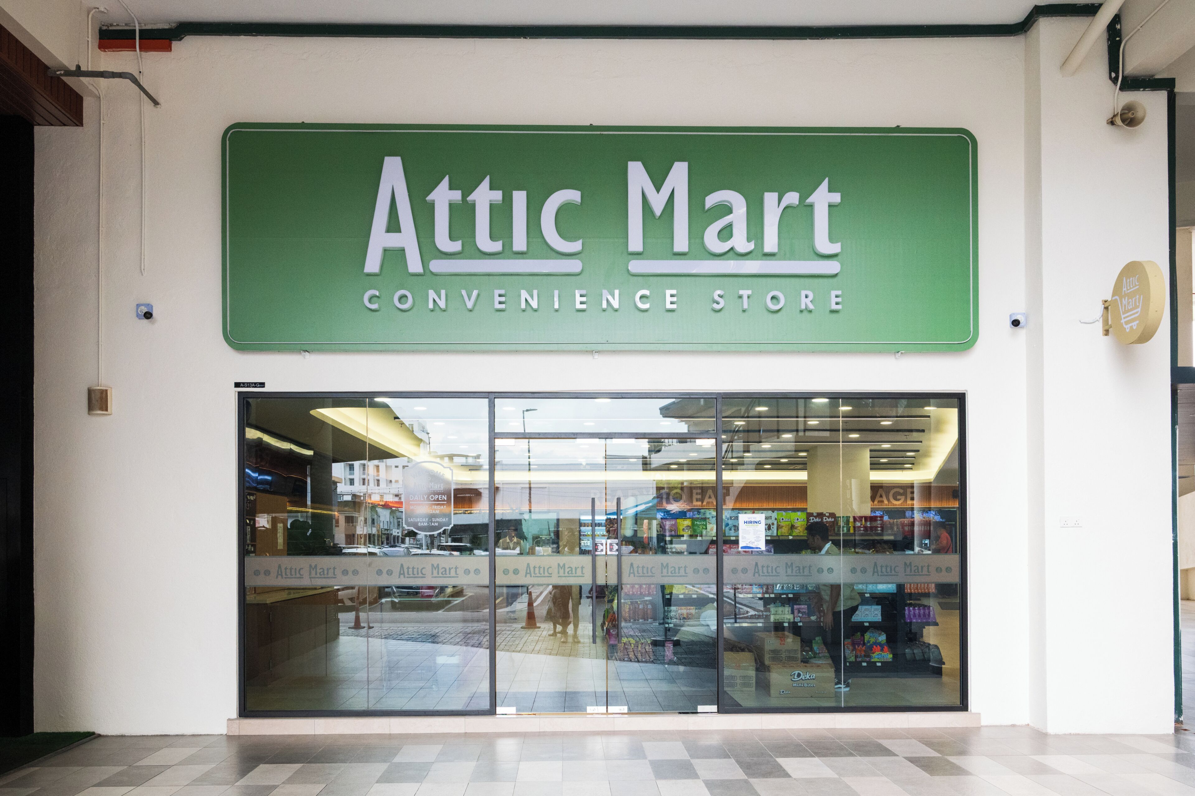 Fachada de loja com um letreiro verde que diz 'Attic Mart Convenience Store' e grandes janelas de vidro.