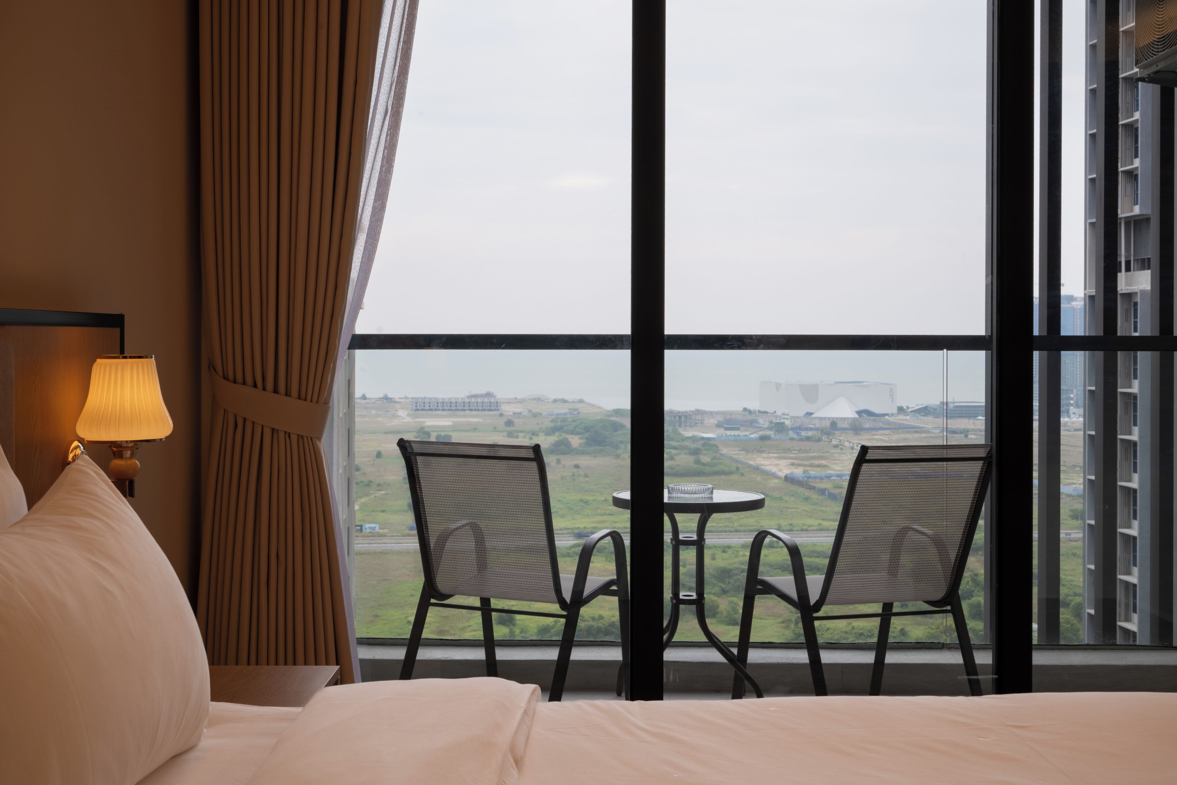 Hotelkamer met een bed, een lamp, een raam met uitzicht op een stadsgezicht en twee stoelen bij het raam.