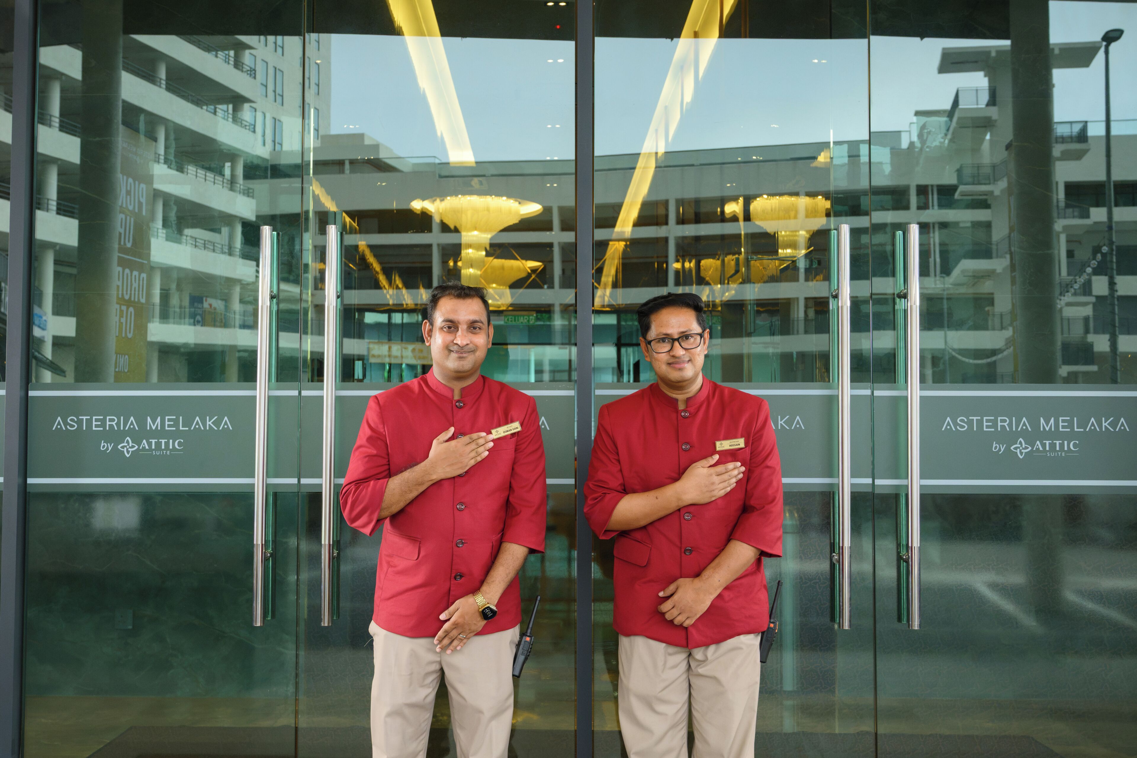 Two individuals standing in front of a building with glass doors, displaying the name 'Asteria Melaka by Attic Suite'.