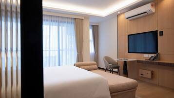 A hotel room with a bed, a desk, a chair, an air conditioning unit, and a television.