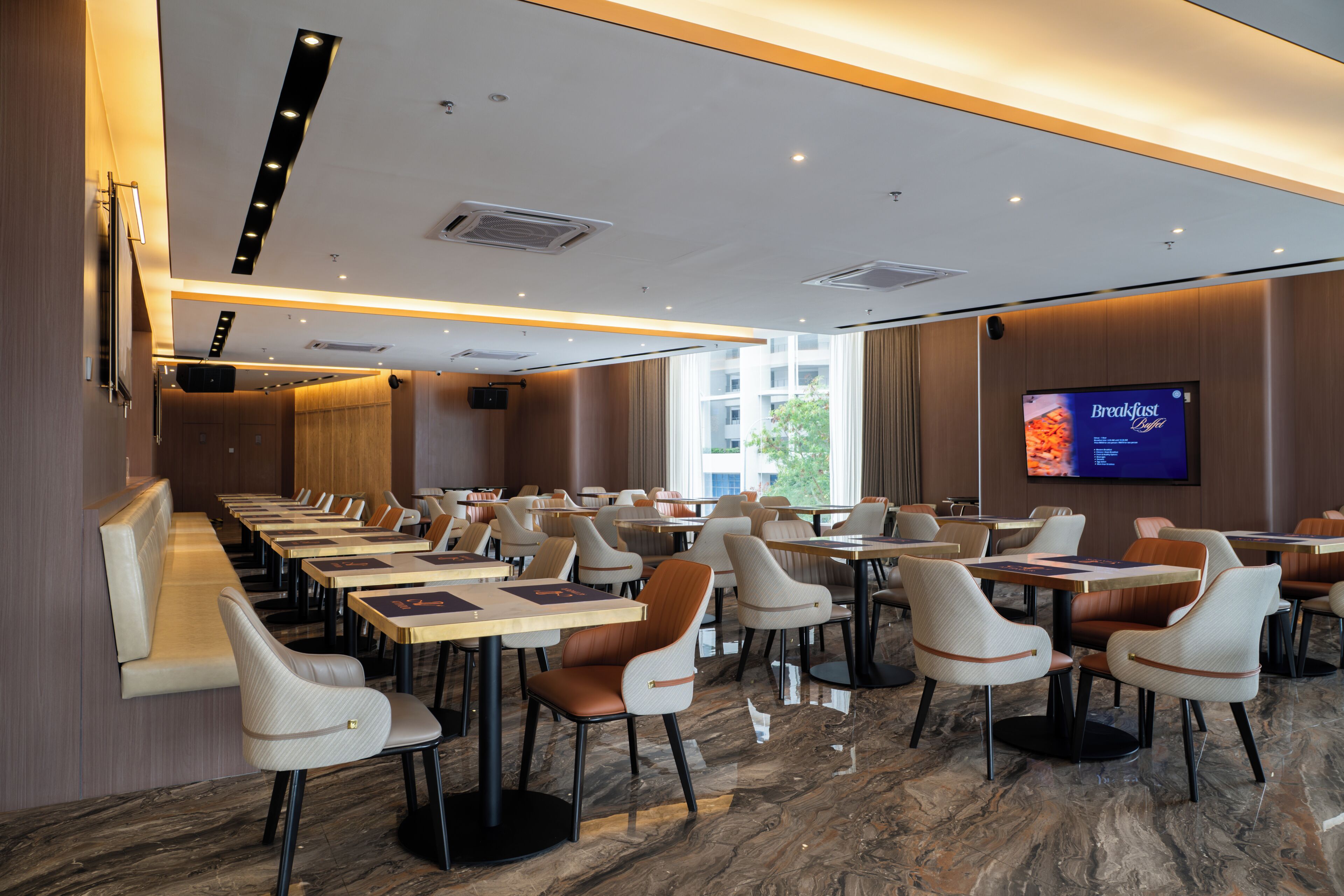 A modern dining area with multiple tables and chairs, a flat-screen TV displaying a breakfast menu, and a large window providing natural light.