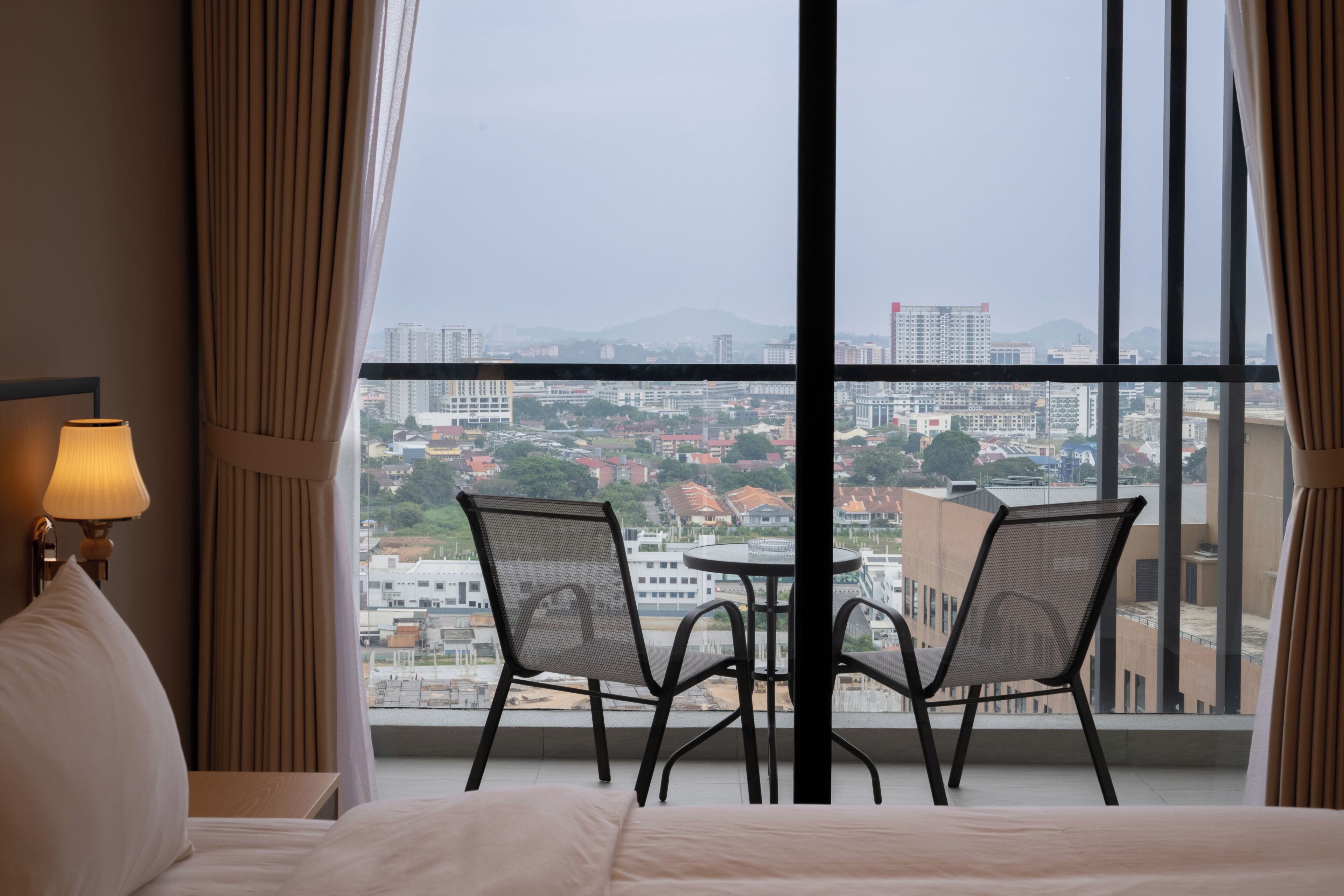 A hotel room with a large window, a bed, two chairs, and a table.
