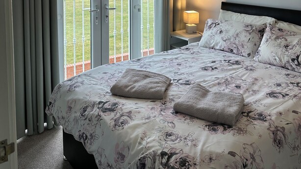 Ein Schlafzimmer mit einem Bett, einem Nachttisch, einer Lampe, einem Gemälde und Blick auf eine grüne Landschaft durch eine Schiebetür.