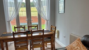 Ein Essbereich mit Holztisch und Stühlen, einer schwarzen Ledersofa und Blick auf ein grünes Feld durch große Fenster mit durchsichtigen Vorhängen.