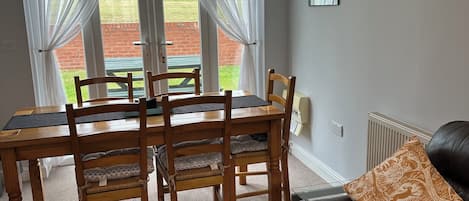 A dining area with a wooden table and chairs, a black leather sofa, and a view of a grassy field through large windows with sheer curtains.
