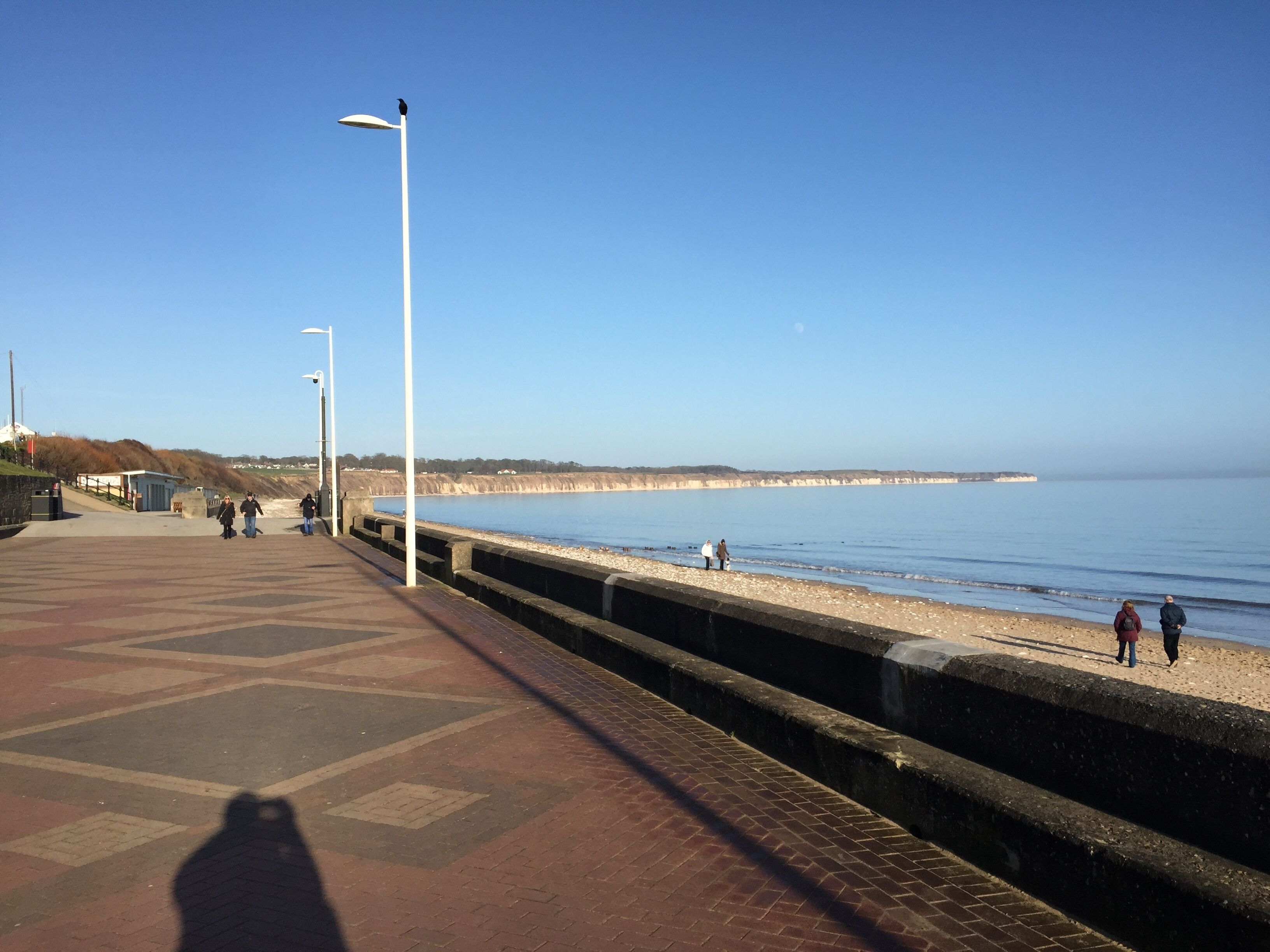 En strandpromenad med fri utsikt över havet, en stenmur och några personer som promenerar.
