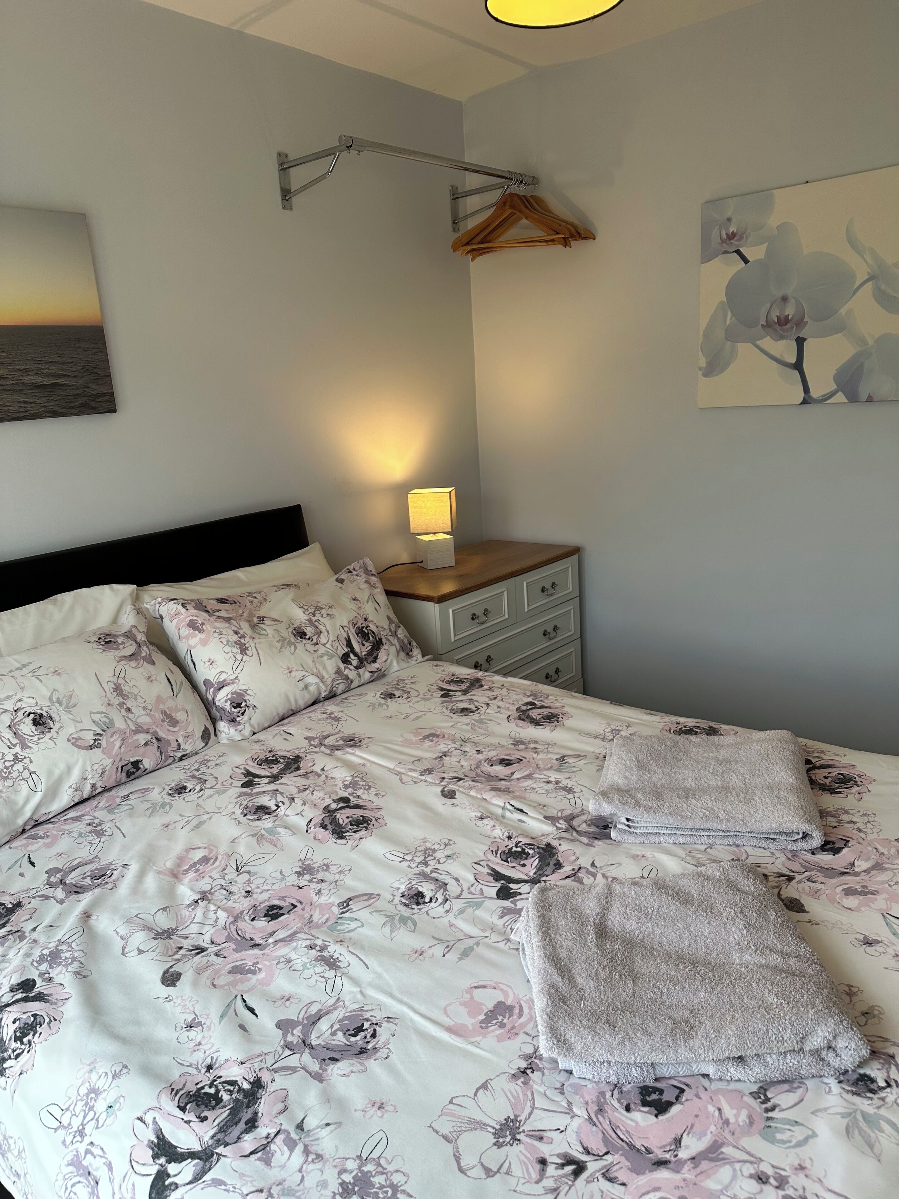 A bedroom with a floral bedspread, a bedside lamp, a wooden nightstand, and a wall-mounted coat rack.