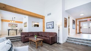 A spacious living area with a brown leather sofa, a wooden coffee table, and a white armchair. A kitchen is visible in the background with a dining table and chairs. - The Mountain House - Walk to the Village! (The Blue Mountains)