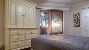 Un dormitorio con cama, cómoda blanca, puerta corrediza de vidrio que da a un espacio exterior y una obra de arte colgada en la pared.
