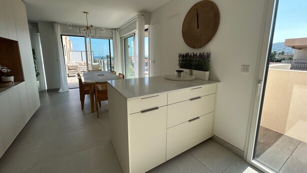 A modern kitchen with a large island, a dining area with a table and chairs, and a view of the outdoors through large windows. - Voiles Blanches 133 by Avecoeur (Vallauris)