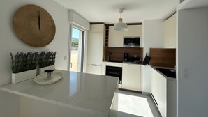 A modern kitchen with a central island, white cabinetry, a round wall clock, and a view of the outdoors. - Voiles Blanches 133 by Avecoeur (Vallauris)