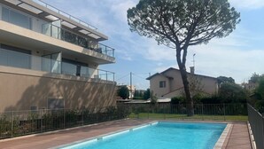 A swimming pool with a wooden deck, a modern building with balconies, and a large tree in the background. - Voiles Blanches 133 by Avecoeur (Vallauris)