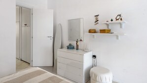 Une chambre à coucher avec une commode blanche, une table de chevet, des toilettes blanches, un miroir et une étagère ornée d’objets décoratifs.