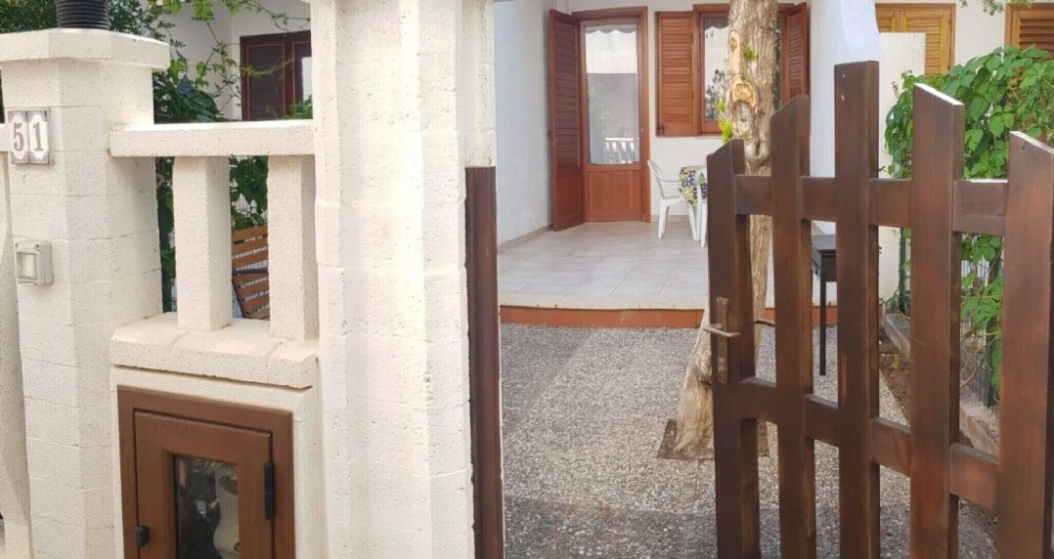 A gated entrance with a wooden door, a white wall with a number sign, and a paved area leading to a building with windows and shutters.