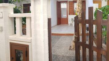 A gated entrance with a wooden door, a white wall with a number sign, and a paved area leading to a building with windows and shutters.
