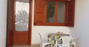 Una habitación con puerta de madera, ventana con contraventanas, mesa con mantel estampado y dos sillas de plástico blancas.