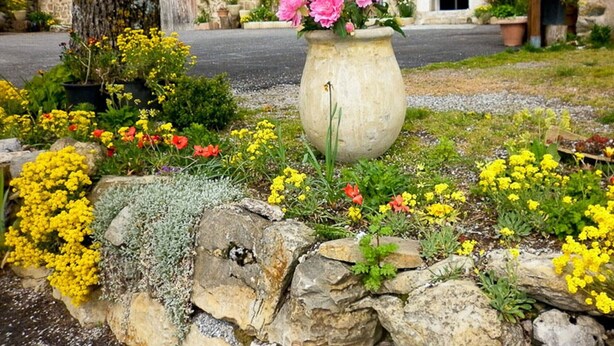 Un edificio in pietra con un giardino antistante, che include un vaso grande pieno di fiori rosa.