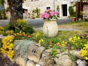 A stone building with a garden in front, featuring a large flower pot with pink flowers.